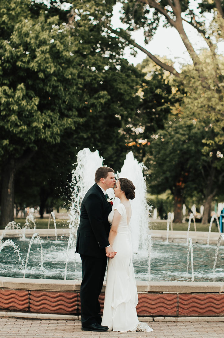 Bride in a crepe fit and flare wedding gown with cap sleeves and a low back, kisses groom by a large outdoor fountain. 