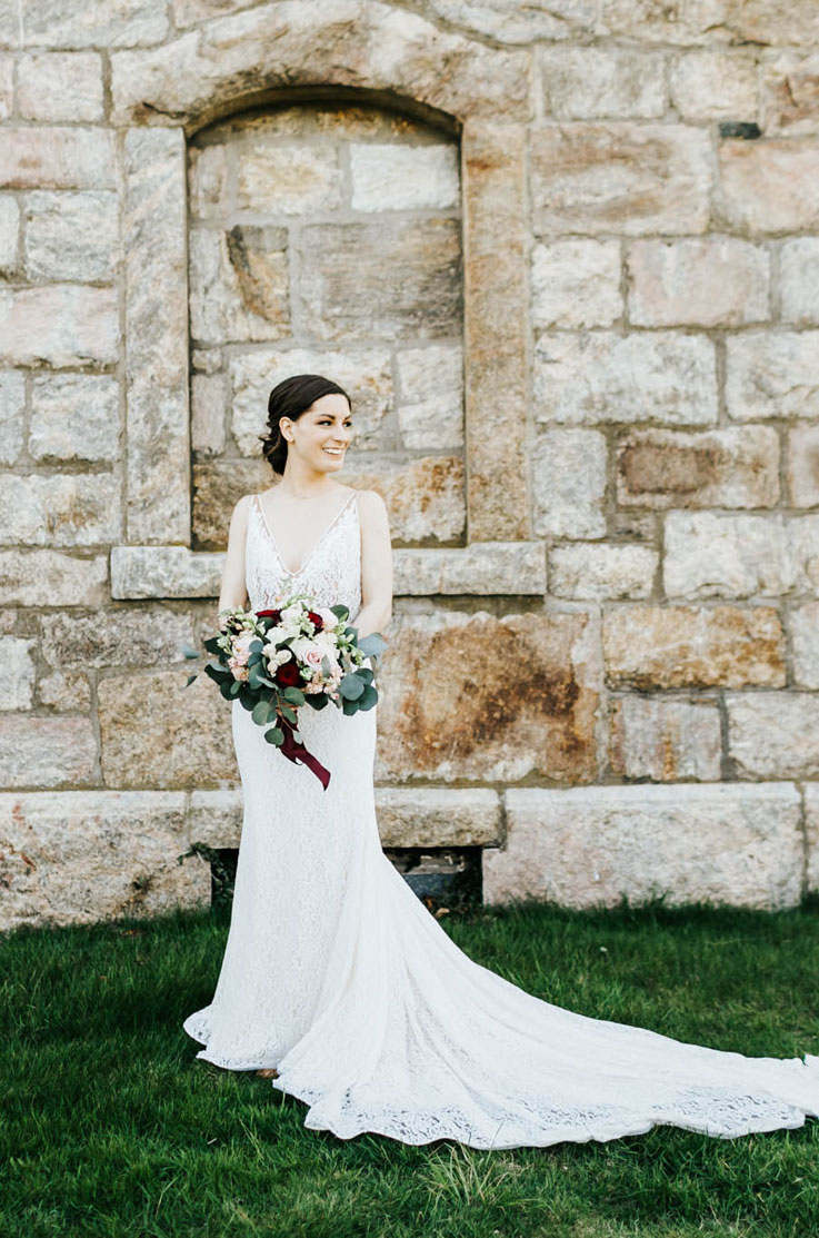 Smiling bride in a lace fit and flare wedding dress  with a sleeveless V-neck bodice, holds a large bouquet by a stone building.