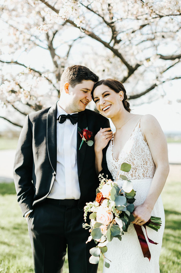 Bride smiles beside groom, wearing a sleeveless lace gown with a V neckline and holds a bouquet under a cherry blossom tree.
