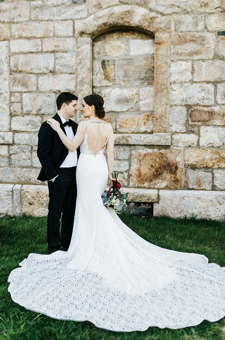 Back of bride in a fit and flare lace gown with a deep-V back and flowing train, embraces groom in a stone wall courtyard.