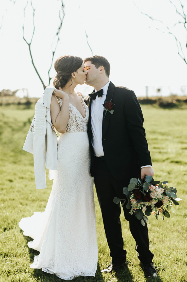 Groom in a black tux kisses his bride in a lace fit and flare gown with a V-neck and is holding a jacket over her shoulder.