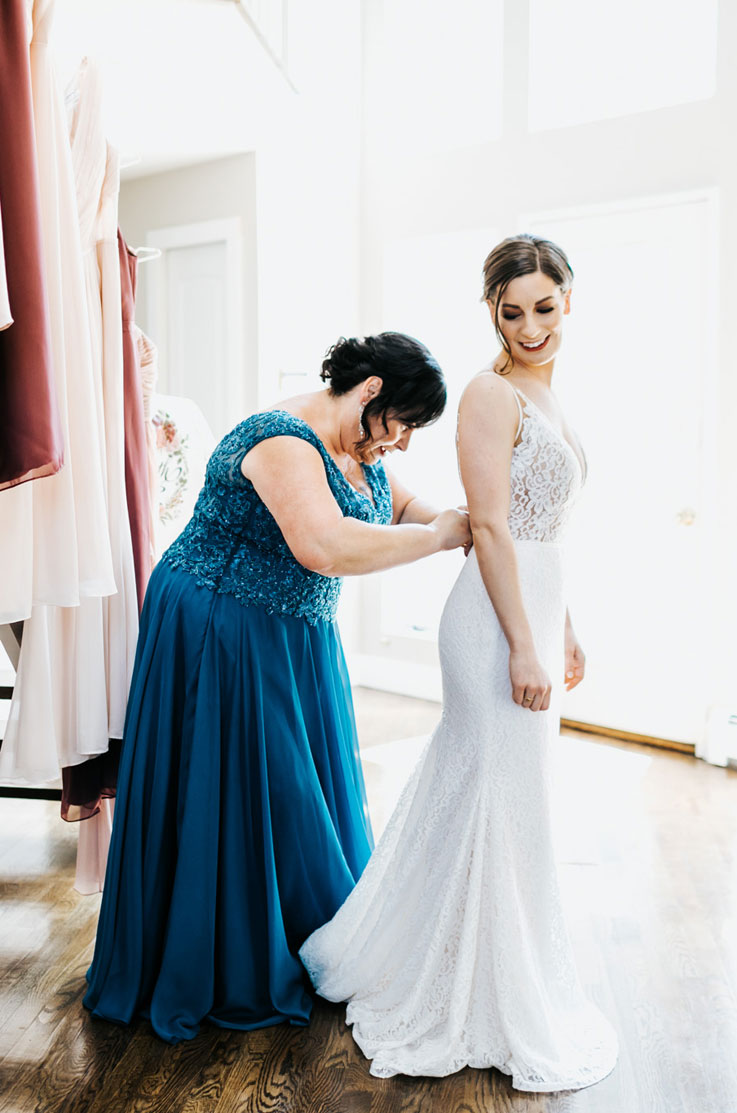 Bride is being fastened into a sleeveless lace gown with a V-neck and is standing indoors by tall bright windows.