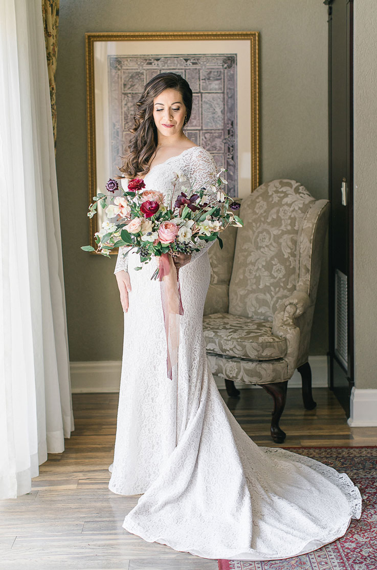 Bride poses in a long-sleeved lace gown with a plunging neckline and chapel train, looking at bouquet by a chair and curtain.