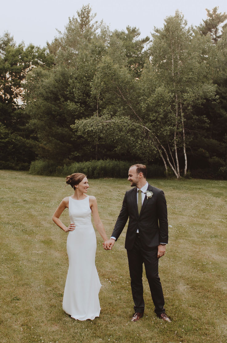 Bride poses beside groom holding hands, in a crepe fit and flare gown with a bateau neck on an open field facing each other.