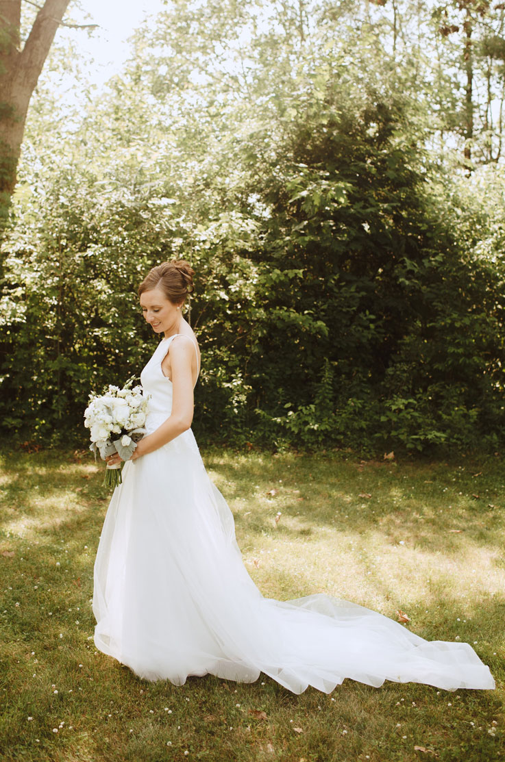 Bride in a sleeveless crepe gown with a bateau neck and overskirt, poses in a sunny forest clearing, holding a white bouquet.