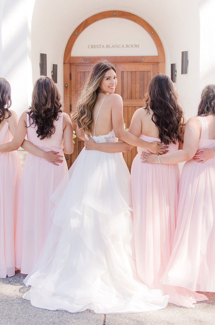 Bride in a multi-layered tulle gown, looking back and stands next to bridesmaids in pink dresses at a Spanish-style venue.