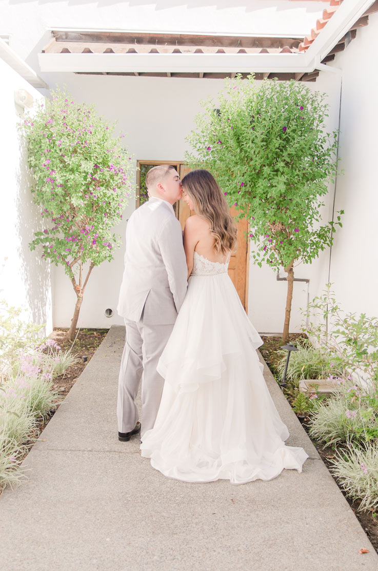Groom kisses his bride on the head, in a lace bodice gown with a layered tulle skirt and thin straps in front of the venue entrance.