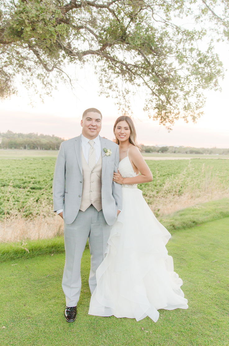  Bride in a spaghetti strap tulle wedding dress poses next to groom, in a gray suit under a tree in front of vineyards.