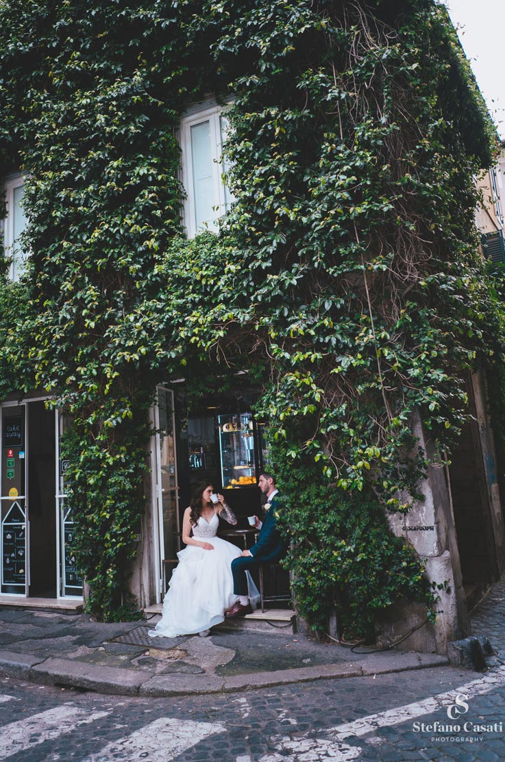 Bride in a layered tulle wedding dress with a V-neckline, sits with groom outside an ivy-covered café on a cobblestone street.