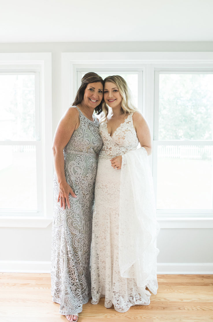 Bride in a lace V-neck wedding dress with tulle and a belt, poses with her mother in brightly lit room with large windows.