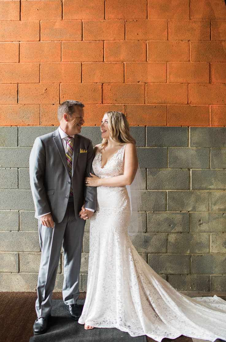 Bride in a sleeveless fit and flare gown with a V-neck lace bodice, smiles next to groom in a gray suit in front of a brick wall.