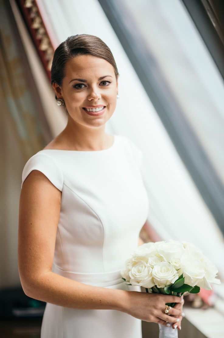 Close-up of bride in a boat neck crepe gown with belt detail and cap sleeves, holding a white rose bouquet by a bright window. 
