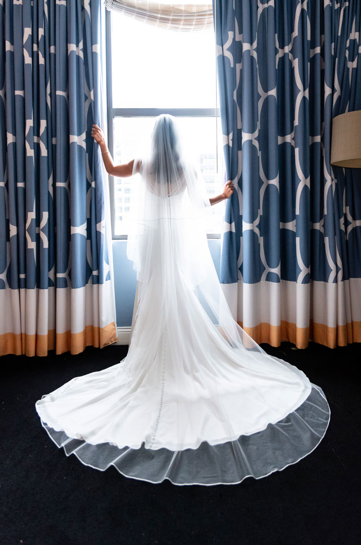 Back of bride in a crepe spaghetti strap gown with a flowing veil and train, poses at a bright hotel window with curtains.