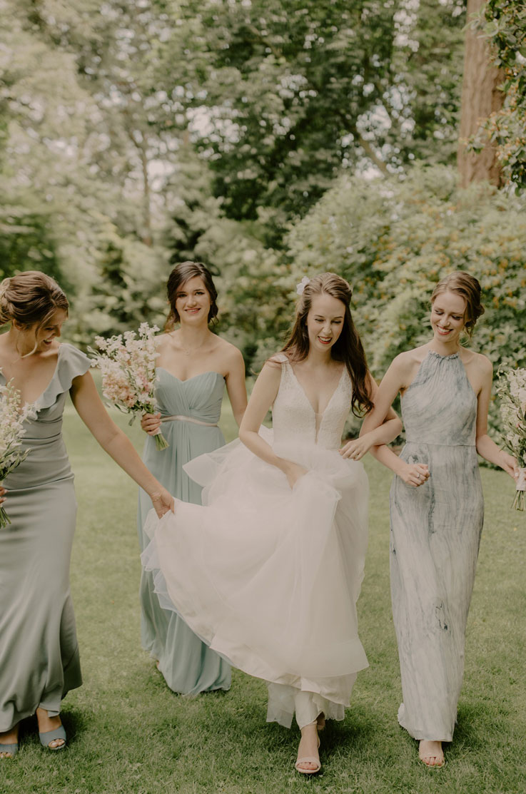 Bride walks smiling in a plunging neck tulle gown with a lace bodice, next to bridesmaids in sage green dresses, past trees.