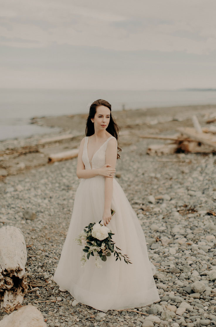 Bride poses in a plunging neck lace gown with a full tulle skirt, and is holding a bouquet on a beachside with driftwood. 