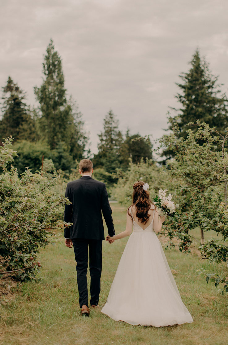 Back of bride in a low open back lace gown with a flowing tulle skirt walking hand in hand with groom through a scenic orchard.