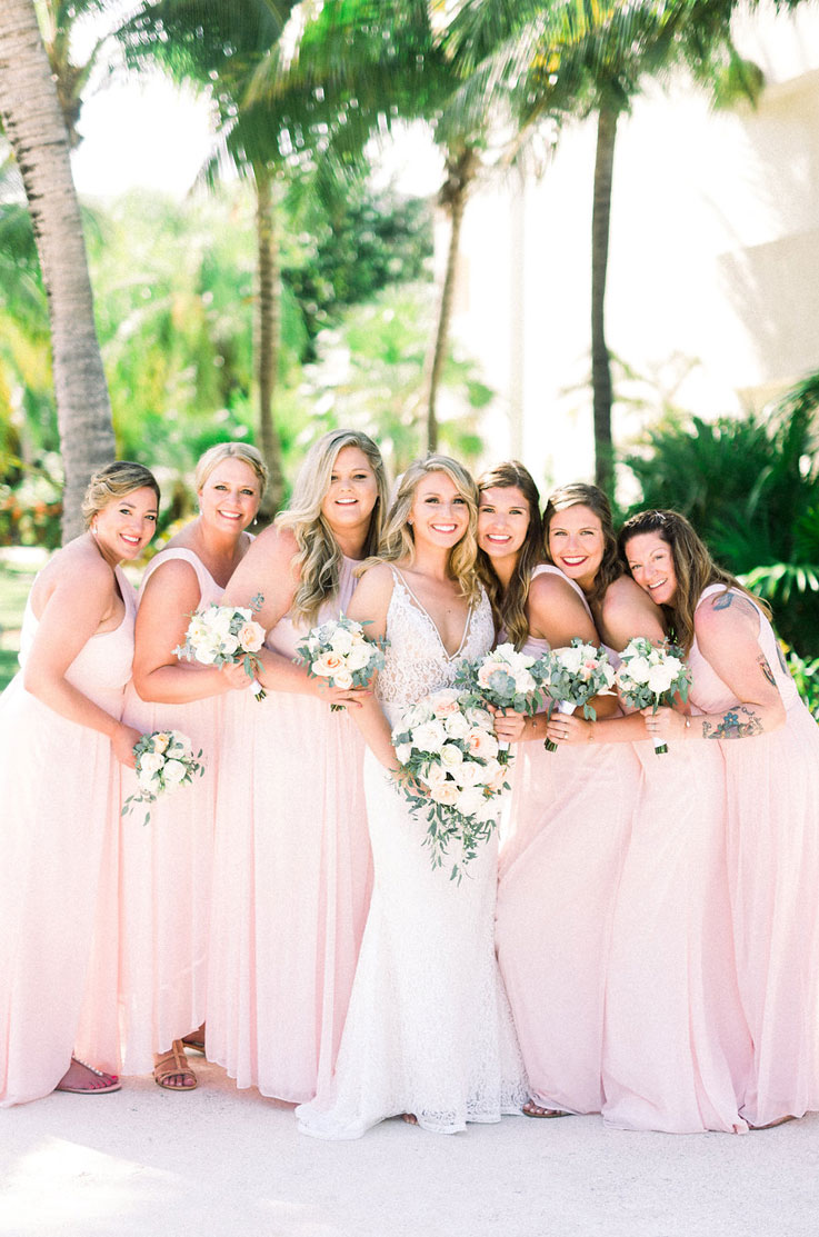 Bride in a lace fit and flare gown with a V-neck, poses with bridesmaids in pink, and are holding bouquets at a resort.