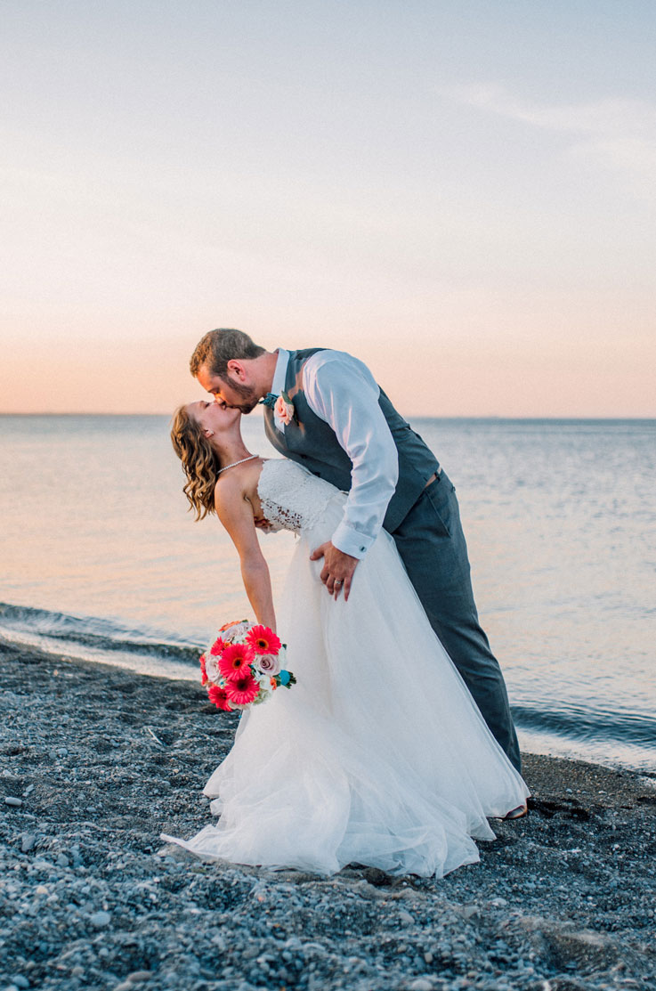 Bride in a strapless Guipure lace bodice wedding dress with flowing tulle and a sweetheart neck, kisses groom on a beachshore.