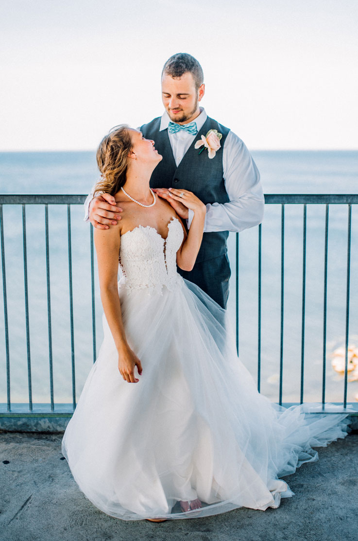 Bride in a guipure lace gown with a tulle skirt, poses with the groom and they face each other on an oceanfront balcony.