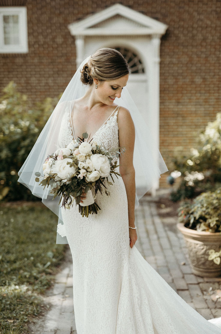 Bride poses in a lace fit and flare gown with a V-neck and veil, holding a bouquet on a stone path outside historic building.