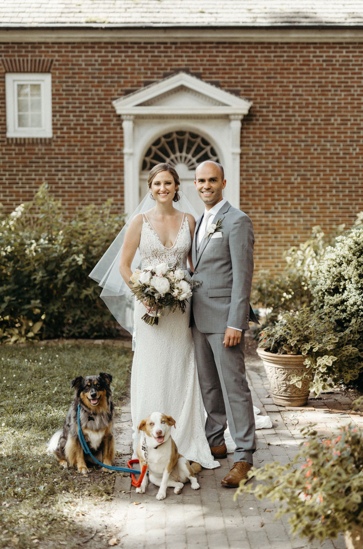 Bride in a V-neck lace gown with a veil, poses next to her groom with two dogs in front of venue entrance near potted plants.