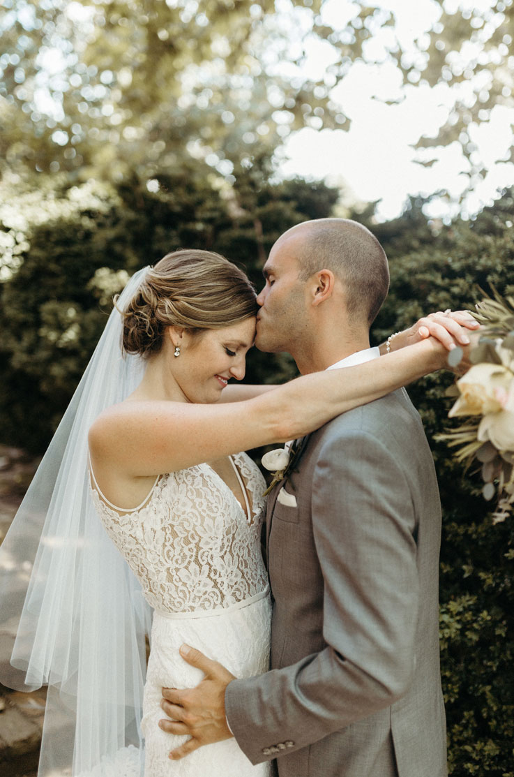 Groom holds the bride in an embrace, wearing a lace bodice gown with a V-neck and veil and is holding a bouquet outdoors.