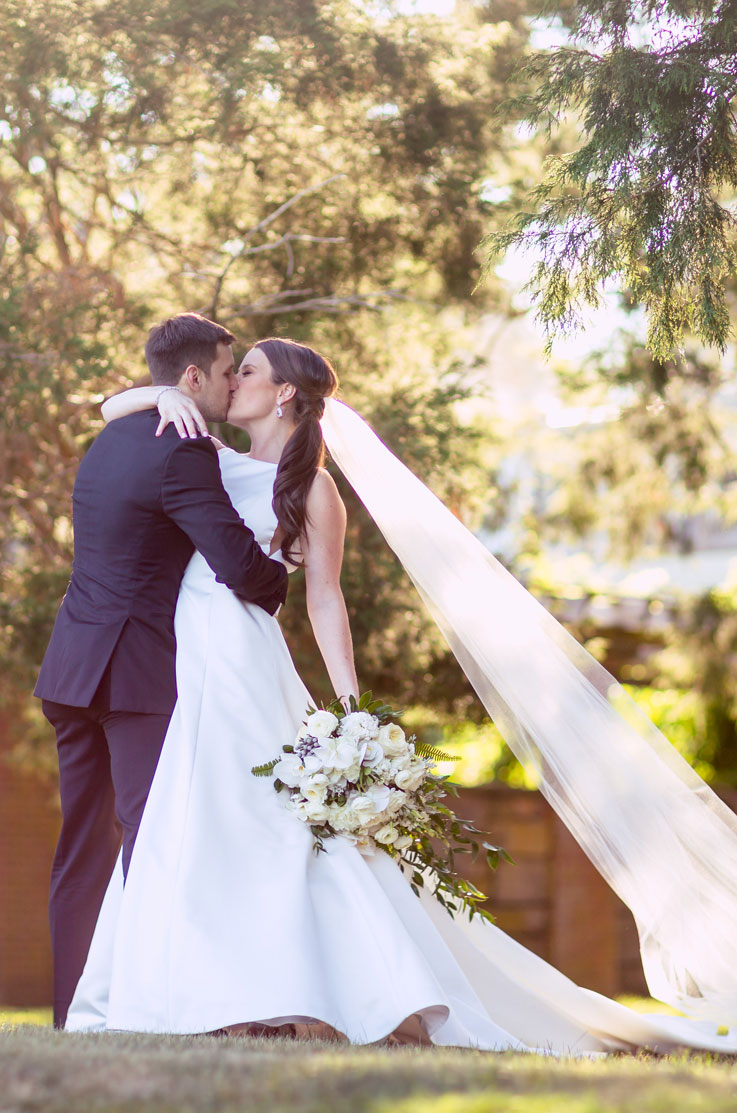 Groom kisses his bride in a satin A-line wedding dress with a bateau neckline and veil, at a sunlit outdoor garden ceremony.