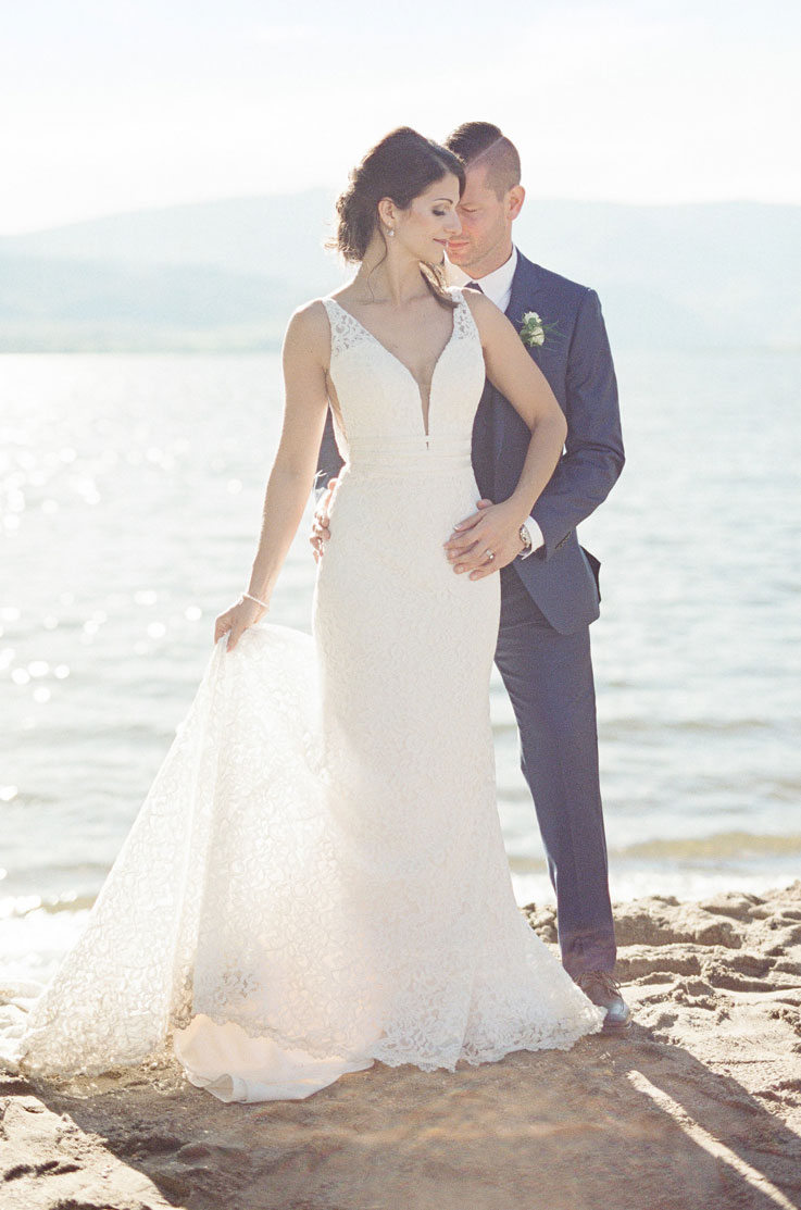 Bride in a lace fit and flare gown with a plunging neck, holding her train and poses with the groom in a blue suit on a beach. 