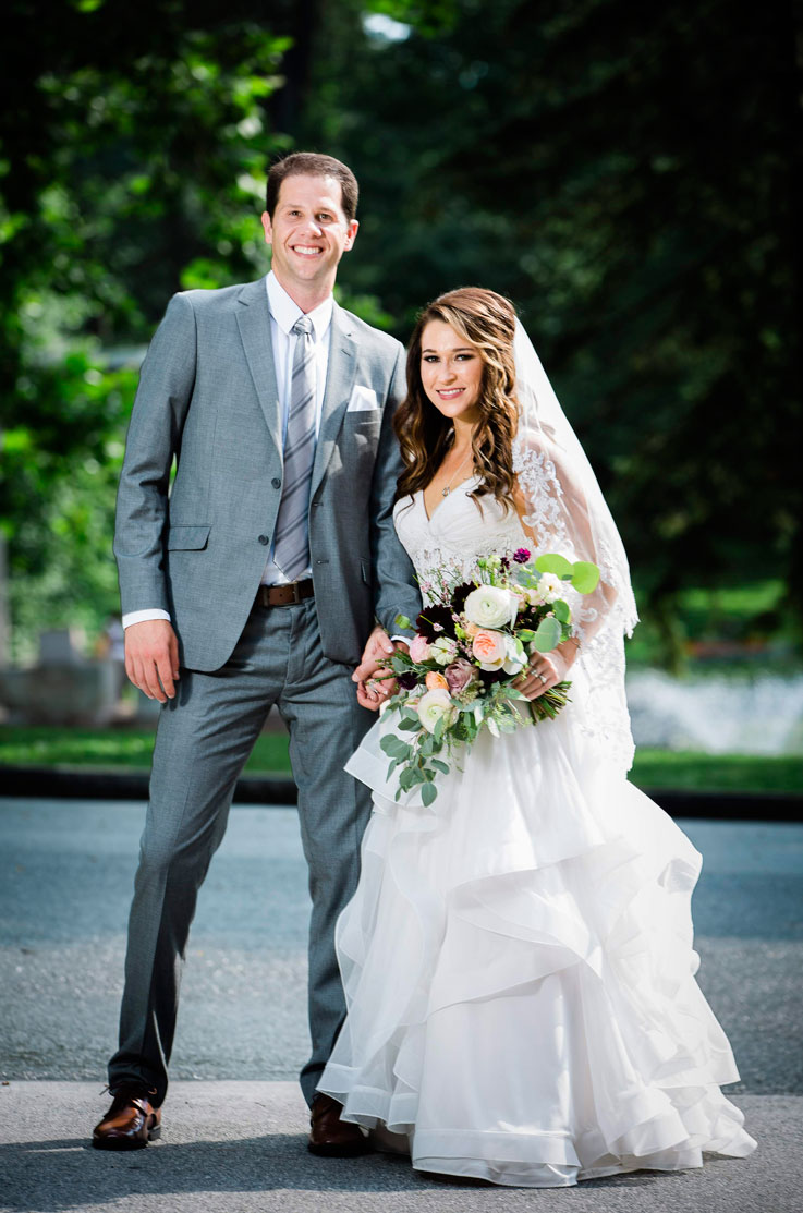Bride in a layered tulle wedding dress with a lace bodice and veil, holds a bouquet and smiles next to groom in gray outdoors.