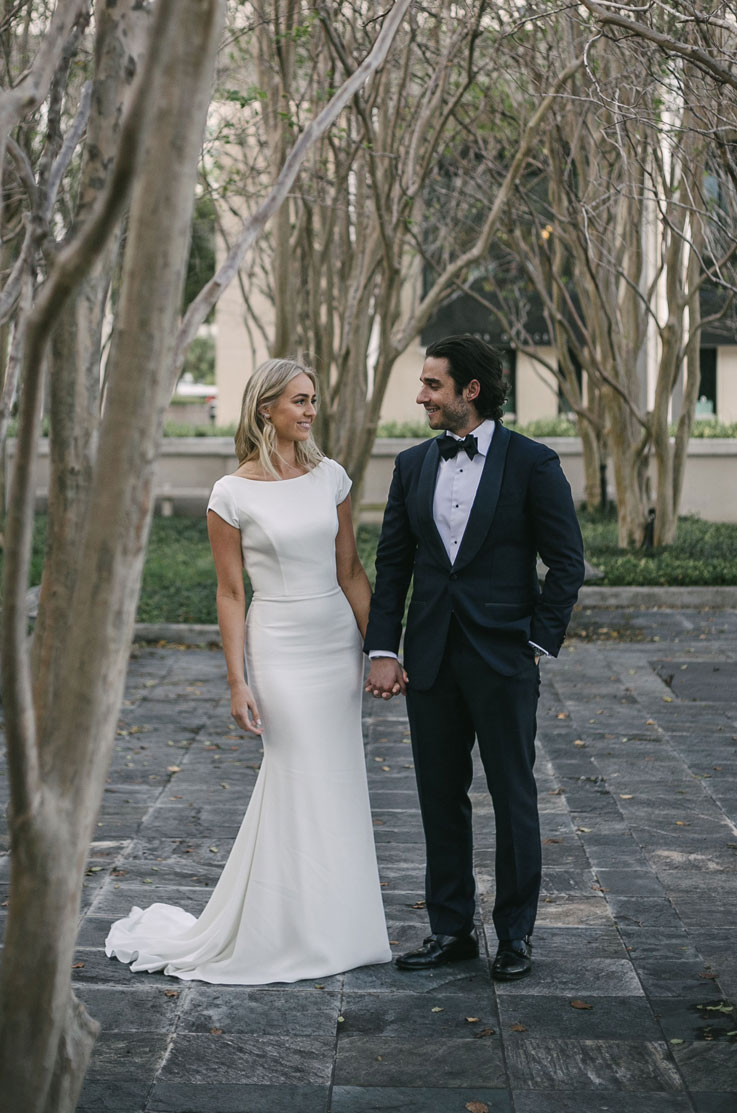Bride in a crepe cap sleeve gown with a chapel train and belt, holds hand with groom as they walk in a tree-lined courtyard.