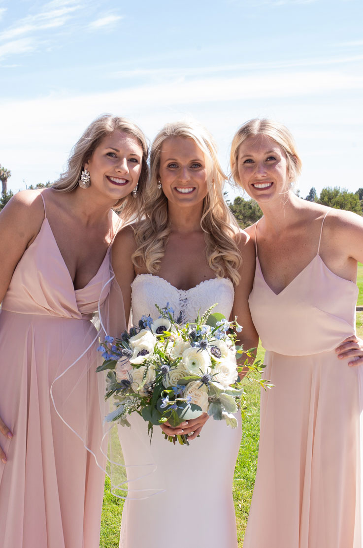 Bride in a strapless lace gown with a crepe fit and flare skirt, holds a bouquet and poses next to bridesmaids in pink dresses.