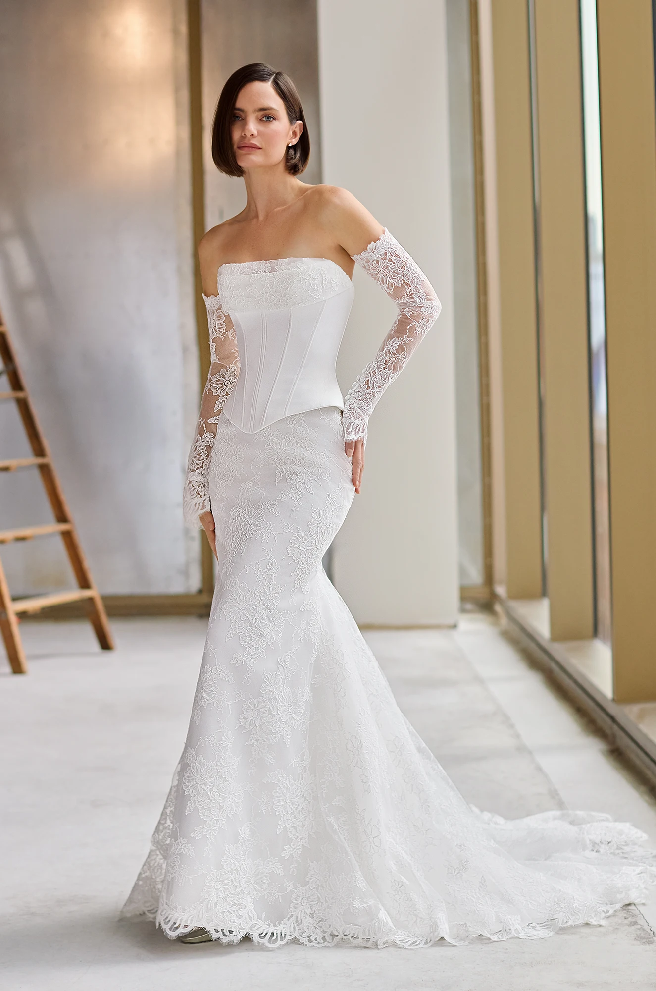 Front view of a strapless Charmeuse wedding dress with corset boning on the bodice, a fitted Lace skirt, and separate Lace sleeves, in a studio with a ladder in the background.