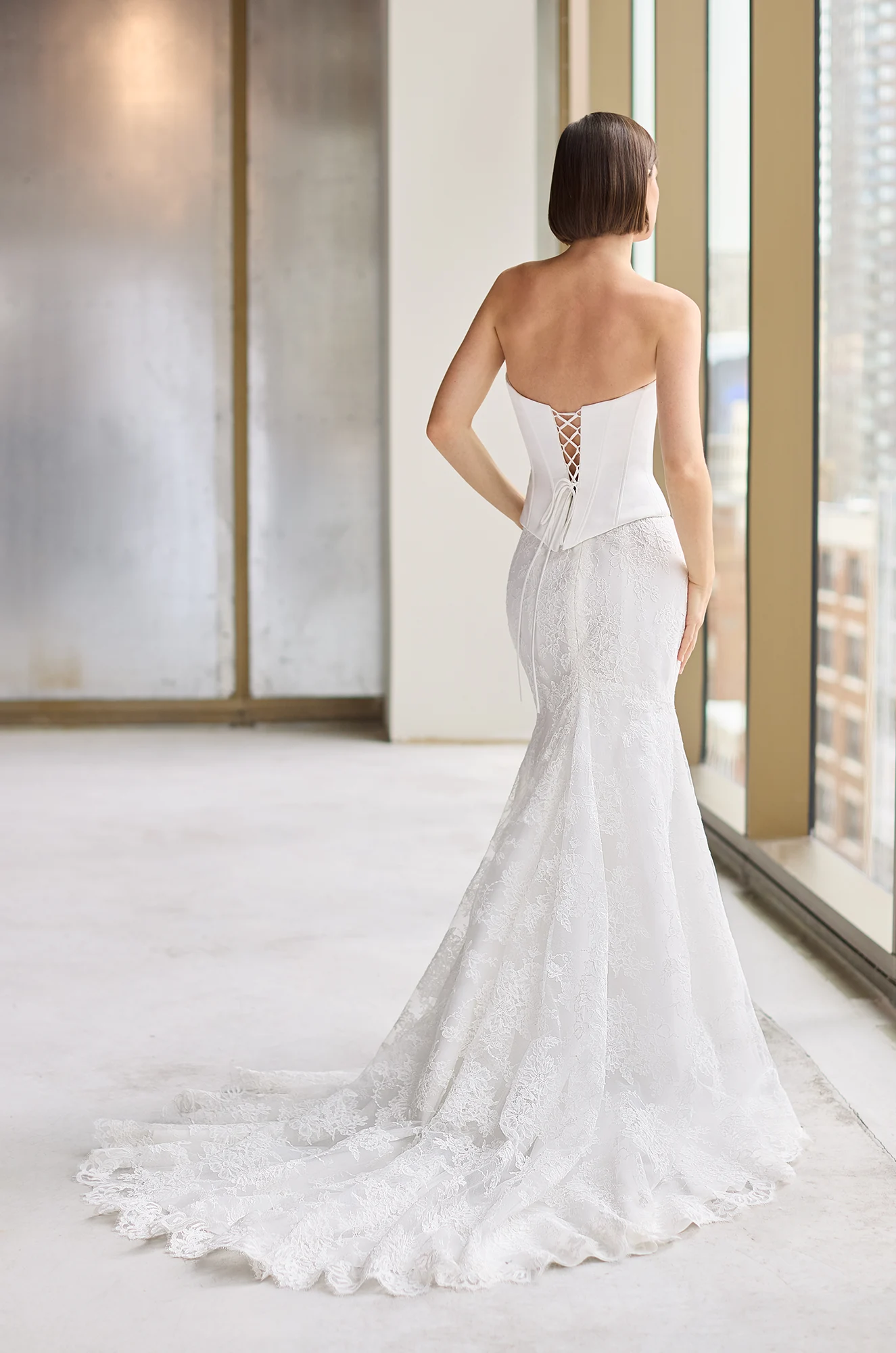 Back view of a strapless Charmeuse wedding dress with corset boning on the bodice and a lace-up back, and a fitted Lace skirt in a studio with large windows.