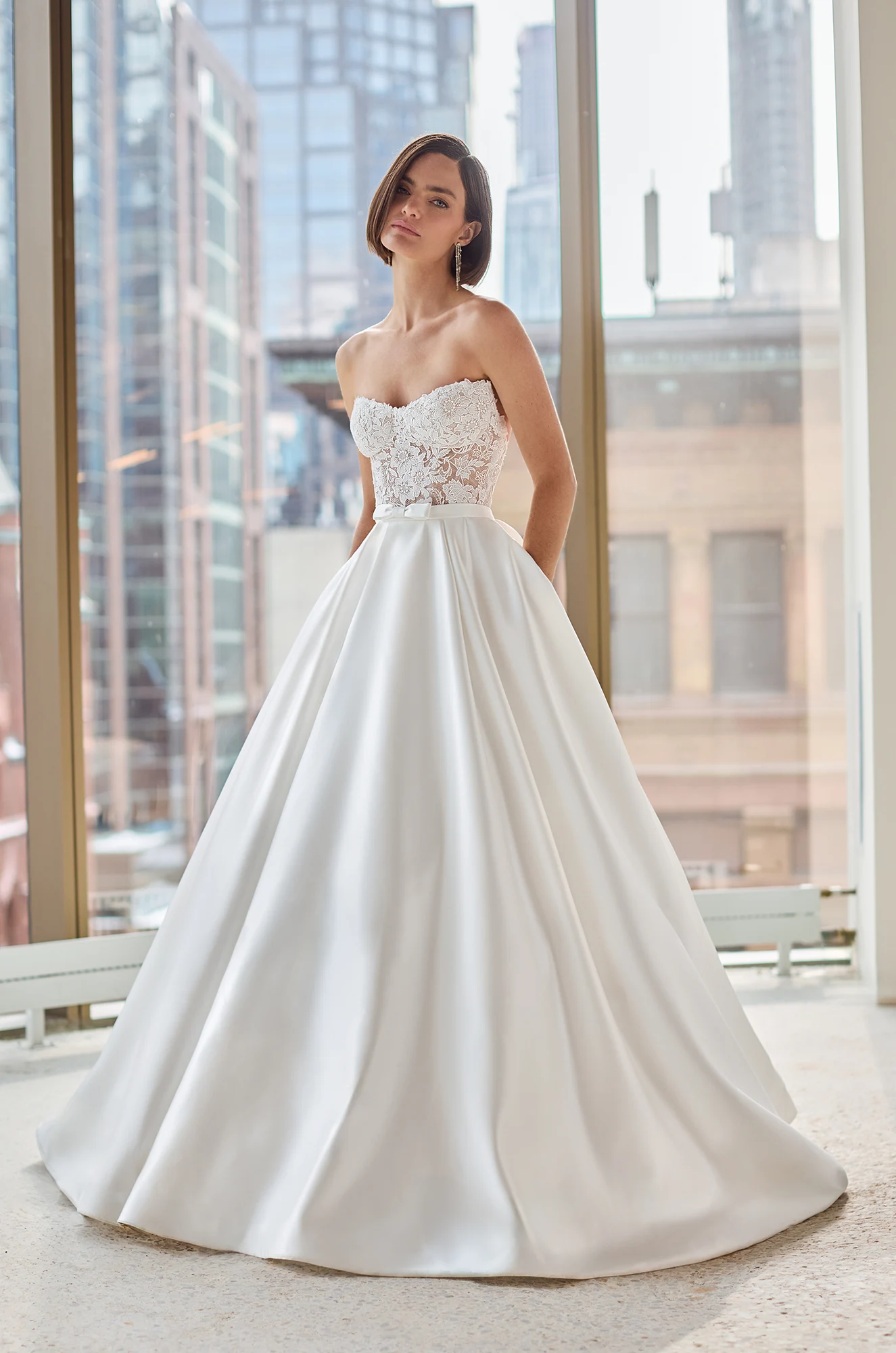 Front view of a strapless lace wedding dress with sheer bodice and Satin ball gown skirt, in front of a large window overlooking a city.
