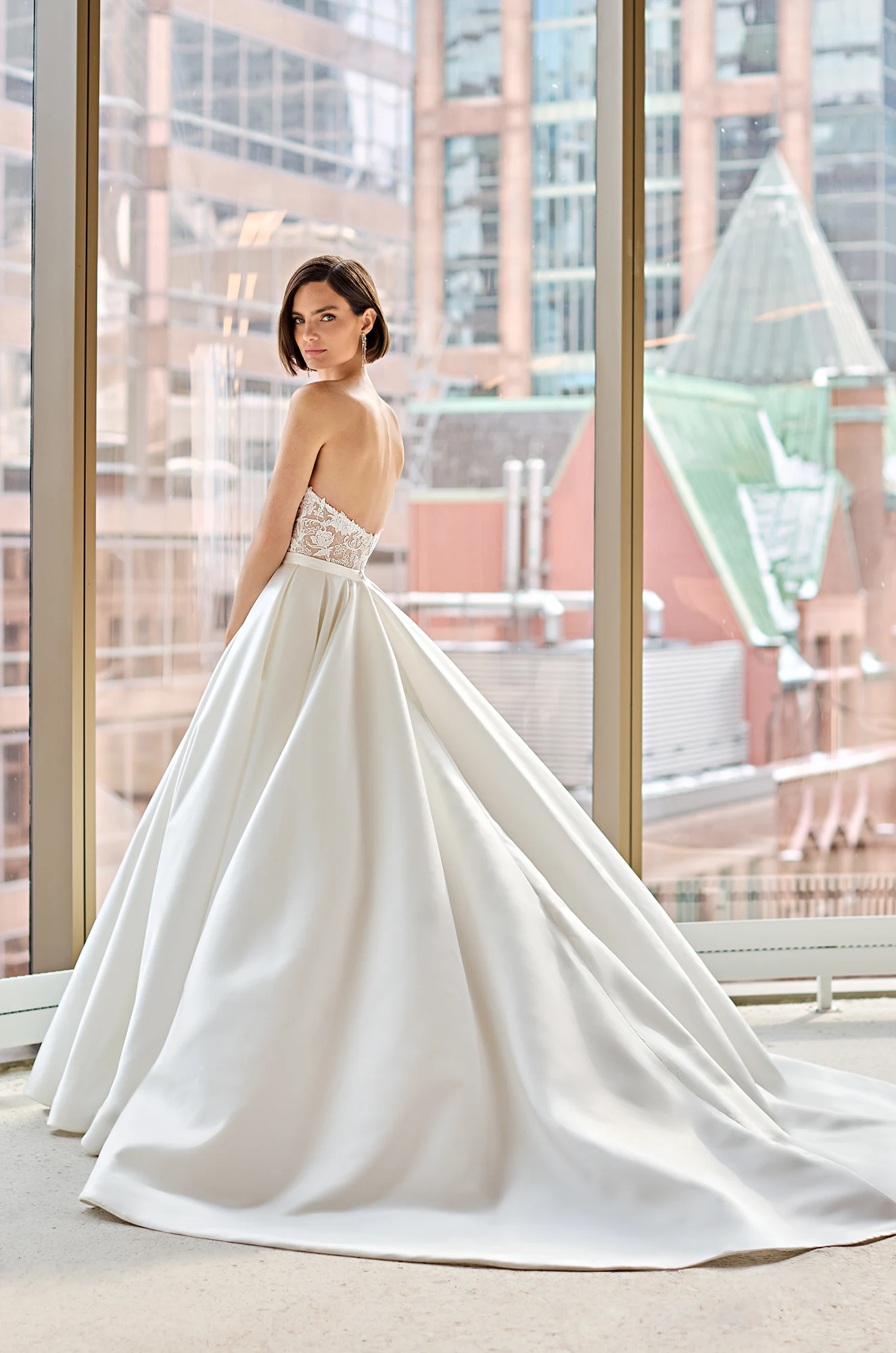 Back view of a strapless Lace wedding dress with sheer bodice and Satin ball gown skirt, in front of a large window overlooking a city.