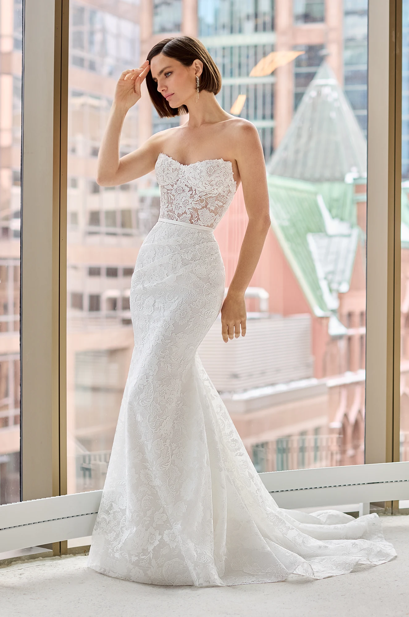 Front view of a strapless Lace wedding dress with sheer bodice and fit and flare lace skirt, in front of a large window overlooking a city.