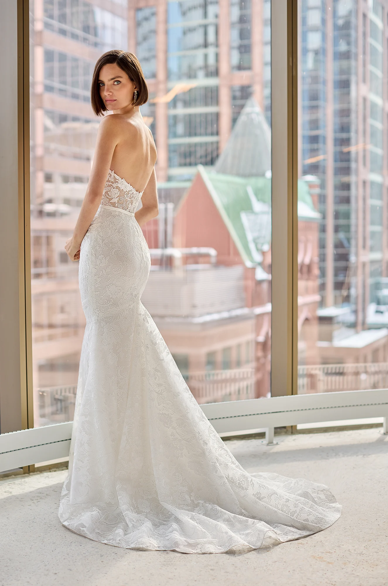 Back view of a strapless Lace wedding dress with sheer bodice and fit and flare lace skirt, in front of a large window overlooking a city.