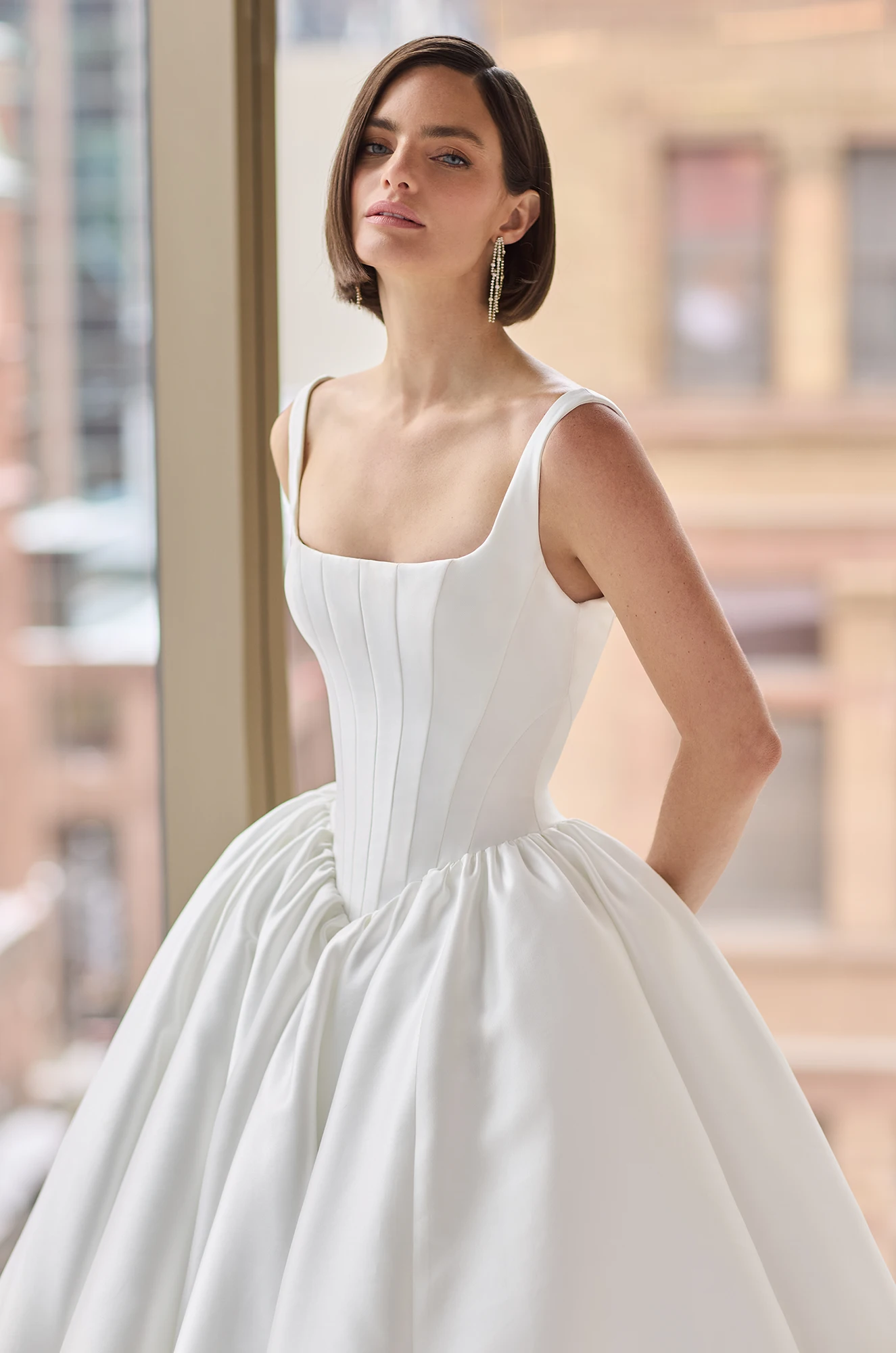 Close up view of a sleeveless Mikado ball gown wedding dress with a seamed bodice and gathered basque waistline, in front of a large window overlooking a city.