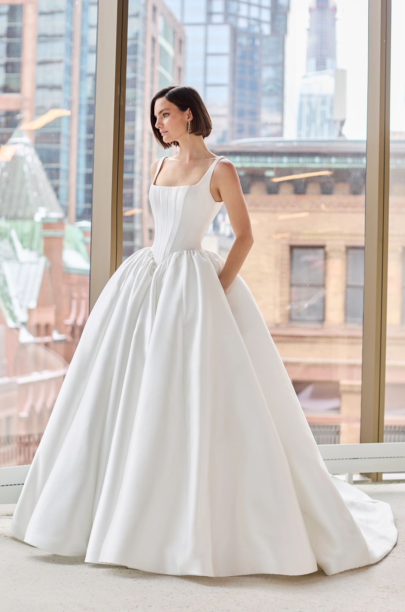 Front view of a sleeveless Mikado ball gown wedding dress with a seamed bodice and gathered basque waistline, in front of a large window overlooking a city. 