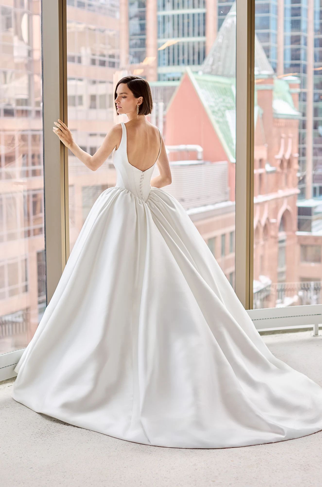 Back view of a sleeveless Mikado ball gown wedding dress with a seamed bodice and gathered basque waistline, in front of a large window overlooking a city.