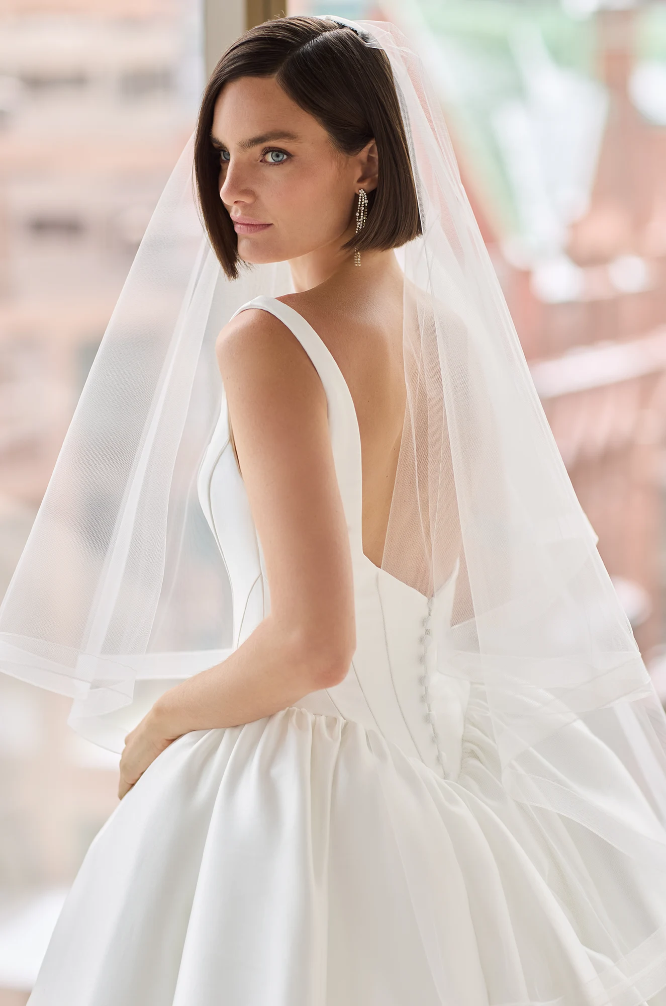 Close up view of a sleeveless Mikado ball gown wedding dress with a seamed bodice and gathered basque waistline, the model is wearing a plain veil and standing in front of a large window overlooking a city.