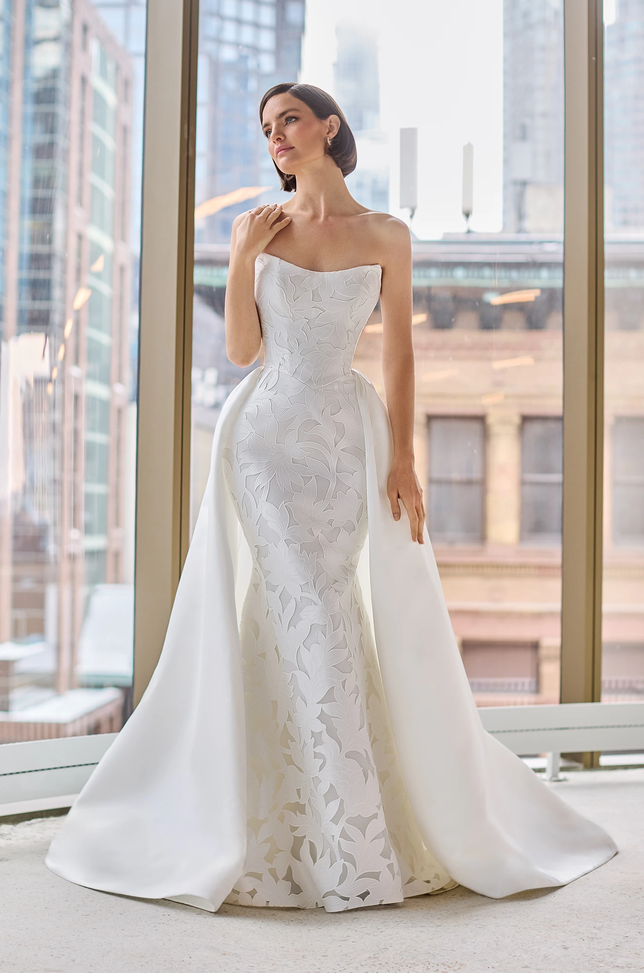 Front view of a strapless Lace wedding dress with a fit and flare Lace skirt and a detachable Satin train, in front of a large window overlooking a city.