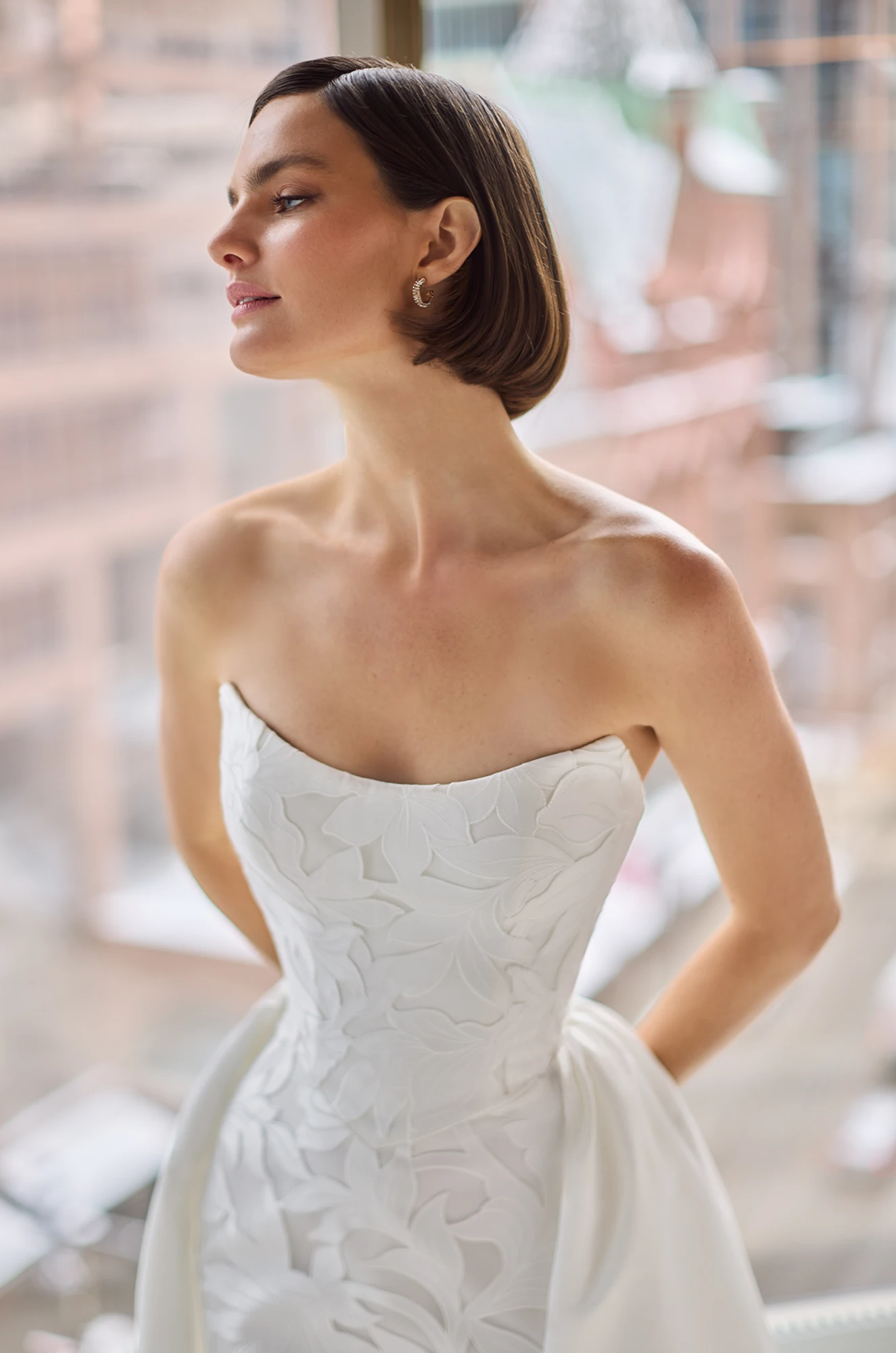 Close up view of a strapless Lace wedding dress with a scoop neckline and a detachable Satin train, in front of a large window overlooking a city.