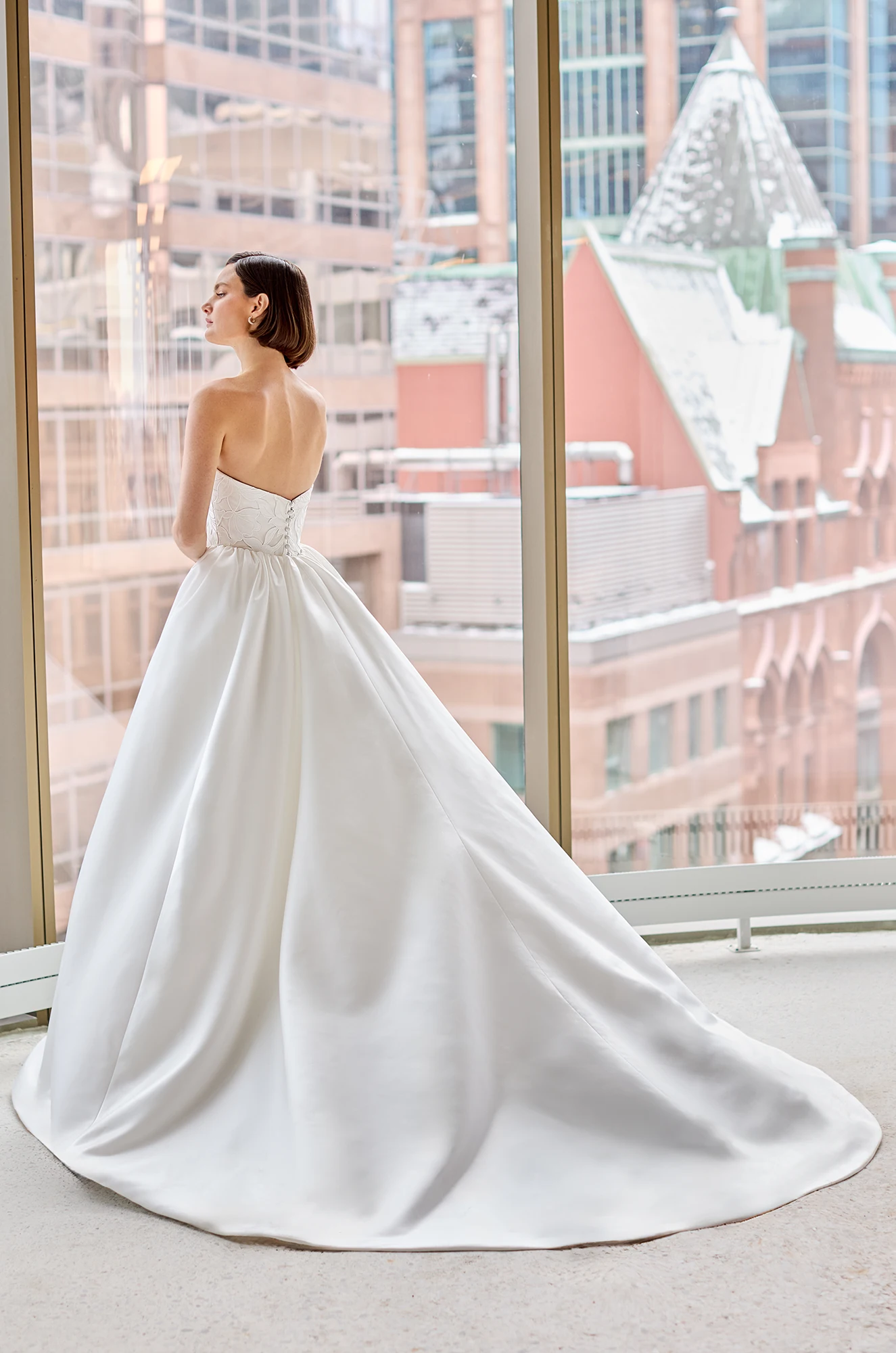 Back view of a strapless Lace wedding dress with a detachable gathered Satin train, in front of a large window overlooking a city.