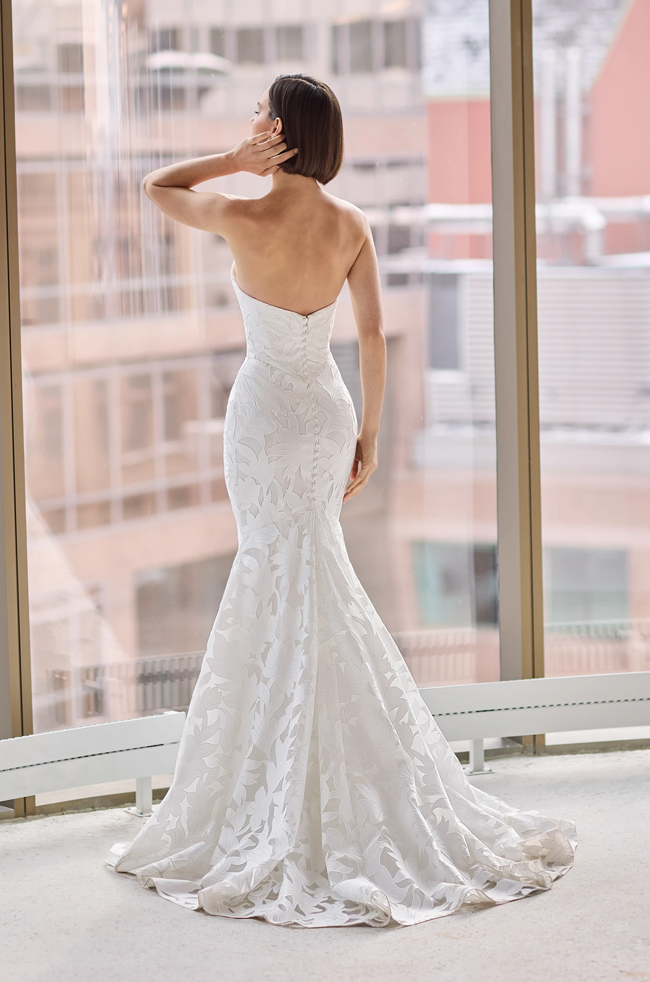 Back view of a strapless Lace wedding dress with a fit and flare skirt, and buttons down the back of the dress, in front of a large window overlooking a city.