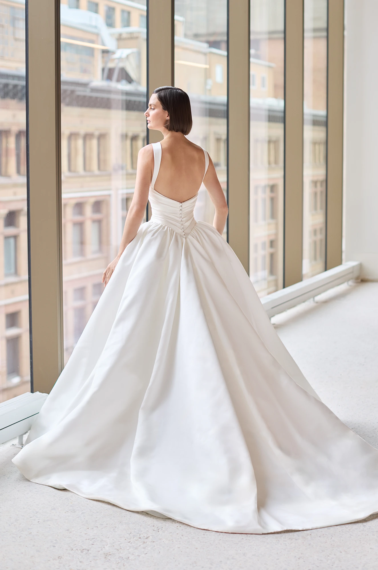 Back view of a sleeveless Satin wedding dress with a draped bodice with buttons to waist, basque waistline, and ball gown skirt, in front of windows overlooking a city.