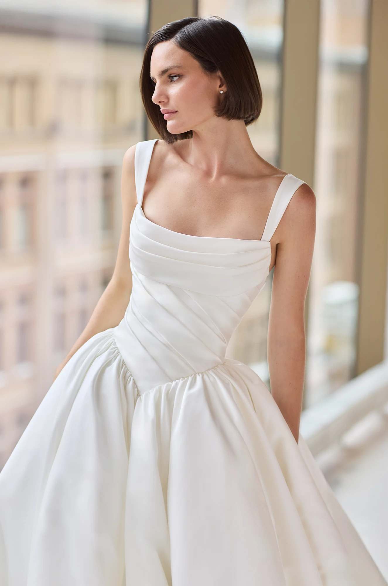Close up view of a sleeveless Satin wedding dress with a draped bodice, basque waistline, and ball gown skirt, in front of windows overlooking a city.