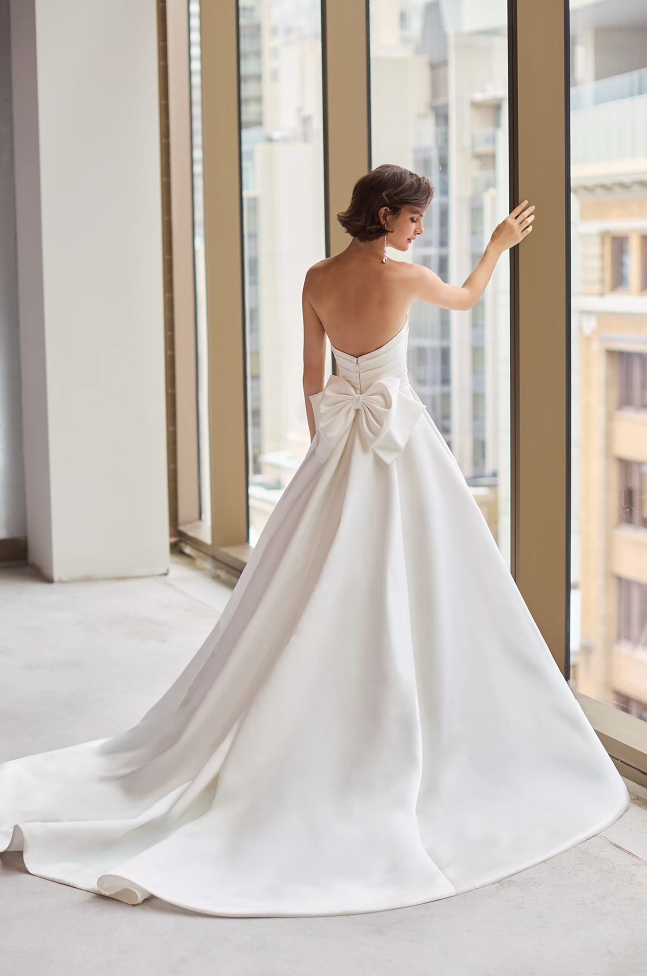 Back view of a strapless Satin wedding dress with a bow on the back of the waist and an A-line skirt, in a studio with large windows overlooking a city.