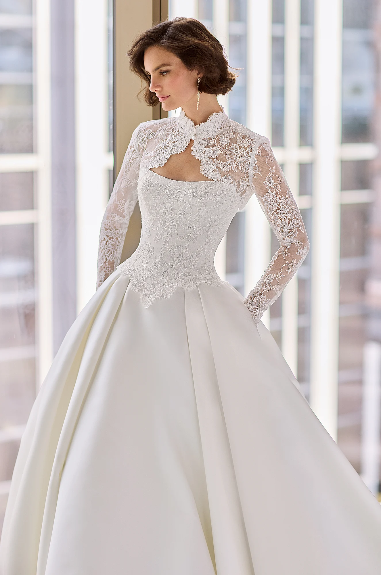 Front view of a strapless wedding dress with a Lace bodice and pleated Satin ball gown skirt, in front of large windows overlooking a city.