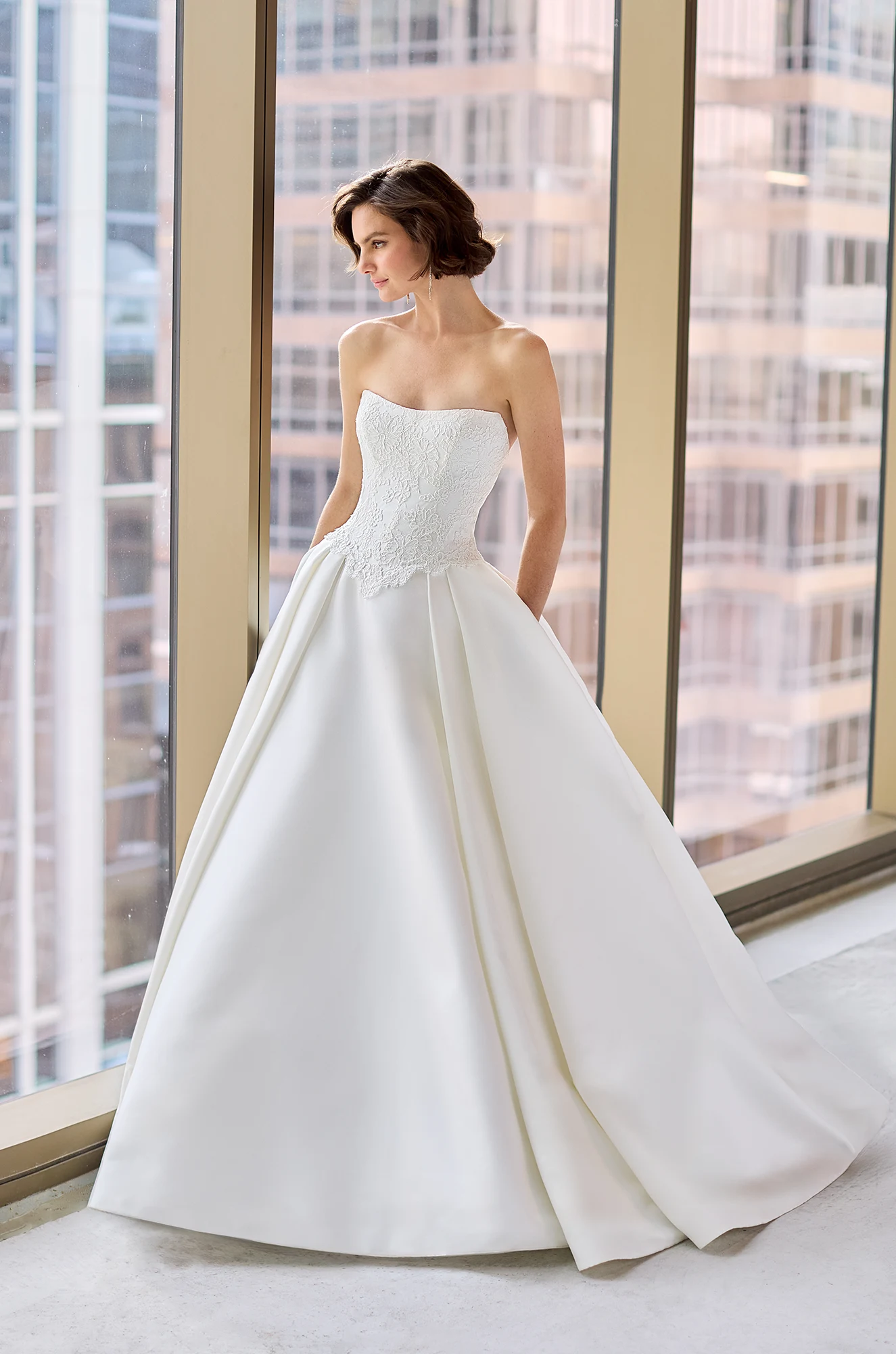 Close up view of a strapless wedding dress with a Lace bodice and pleated Satin ball gown skirt, with a long sleeve Lace bolero, in front of a large window overlooking a city.
